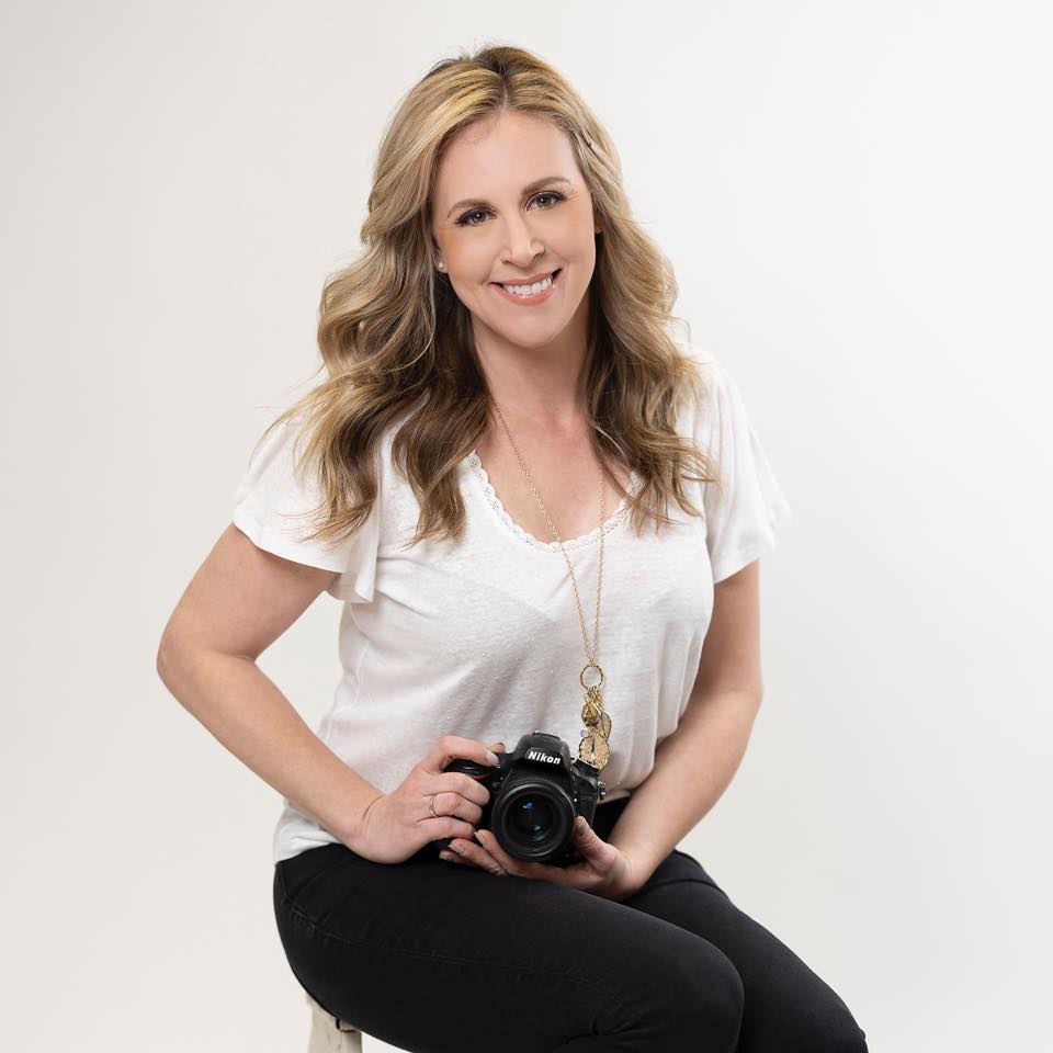 woman with long blonde hair, wearing a white top and black pants, sits on a stool holding a camera. she smiles against a plain white background.